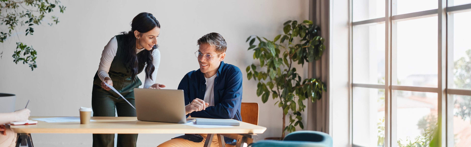 Two colleagues in in coworking setting look at laptop and discuss work while smiling and wearing business casual attire.