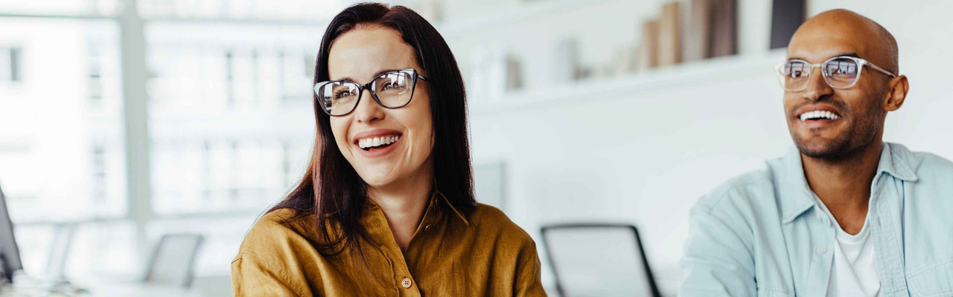 Woman and man both wearing glasses smile at something out of frame, looking to their rights in a brightly lit office setting.
