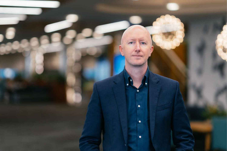 Director of AI and Engineering at Datacom Joel Macfarlane wears navy blazer and shirt in office setting, wit ligts blurred in background, mid distance portrait.