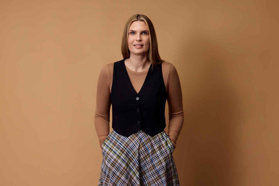 Lou Compagnone Datacom Director of AI wears vest, tan long sleeve and plaid skirt standing in front of neutral background in a mid-distance portrait.