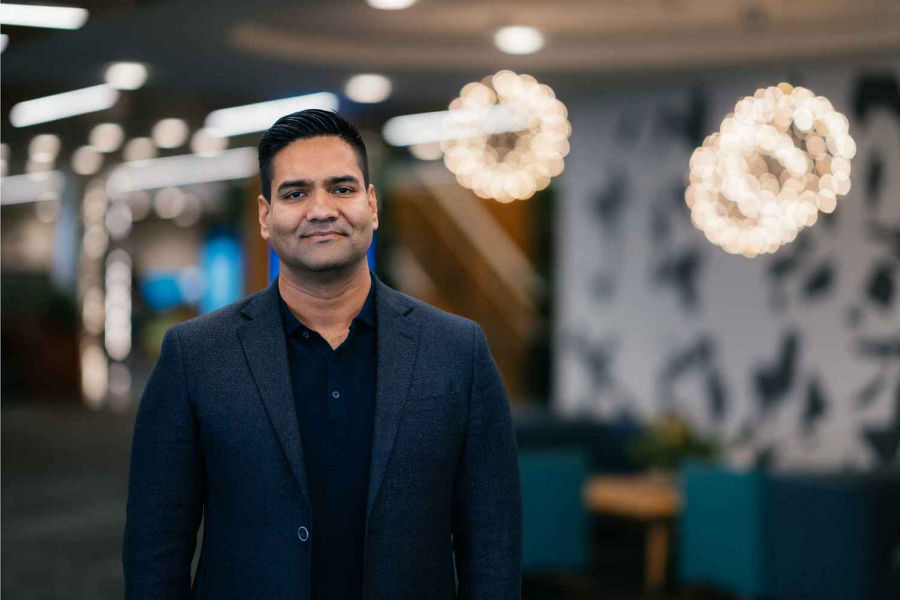 Sunny Katira Datacom Professional Services MD wears navy suit and shirt, smiling in office setting with lights blurred in background in mid-distance portrait.