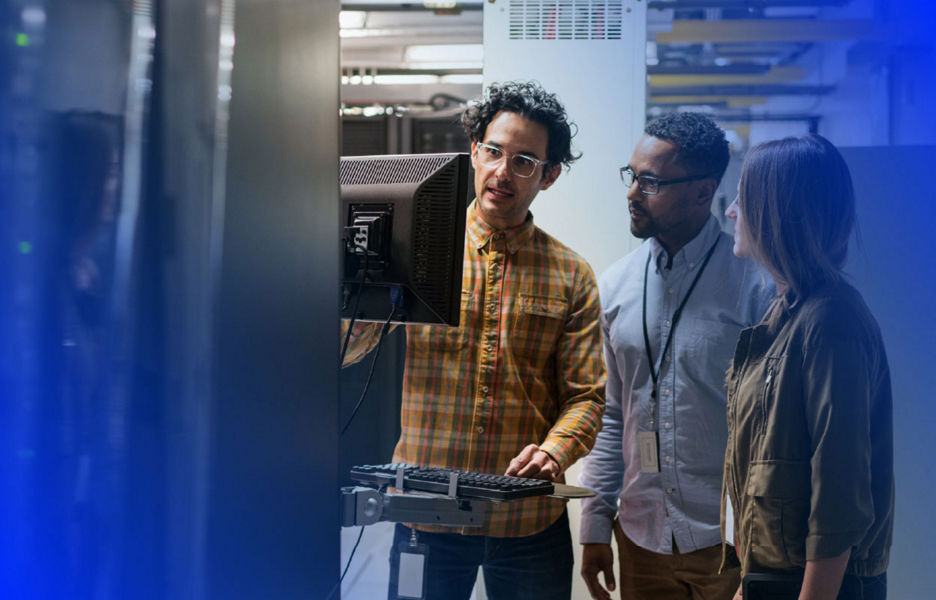 Three individuals looking at a computer screen while standing in a data centre with servers around them