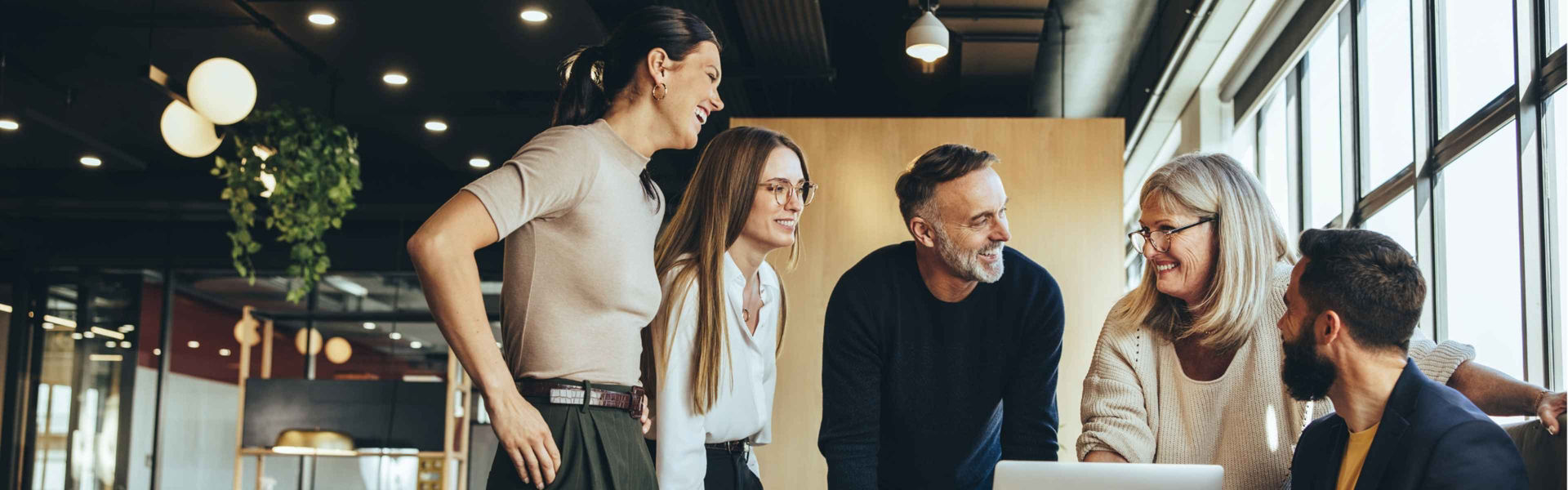 Group of colleagues stand around a computer chatting and smiling in modern office setting.