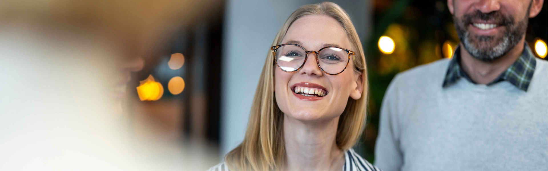 Blonde woman wearing glasses and striped shirt smiles at blurred figure in foreground while colleague stands to her side, office setting with lights in background.