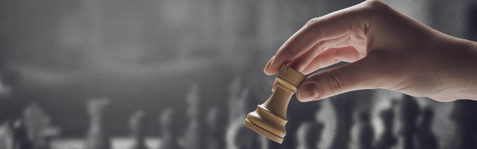 Right hand holding 'rook' chess piece with grey chess board and pieces in the background.