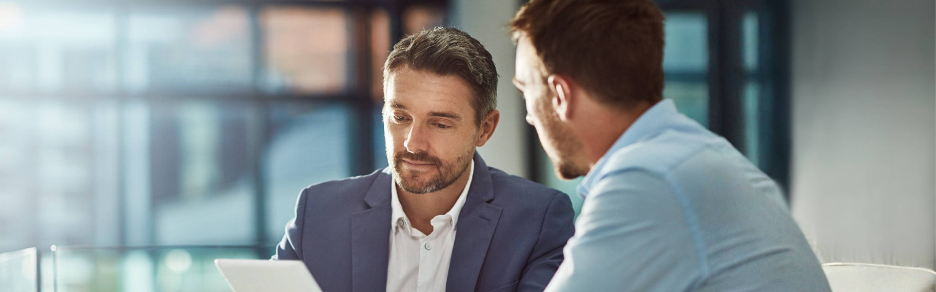 Men sit in business setting discussing and looking at paper, windows and light behind them, wearing formal officewear.