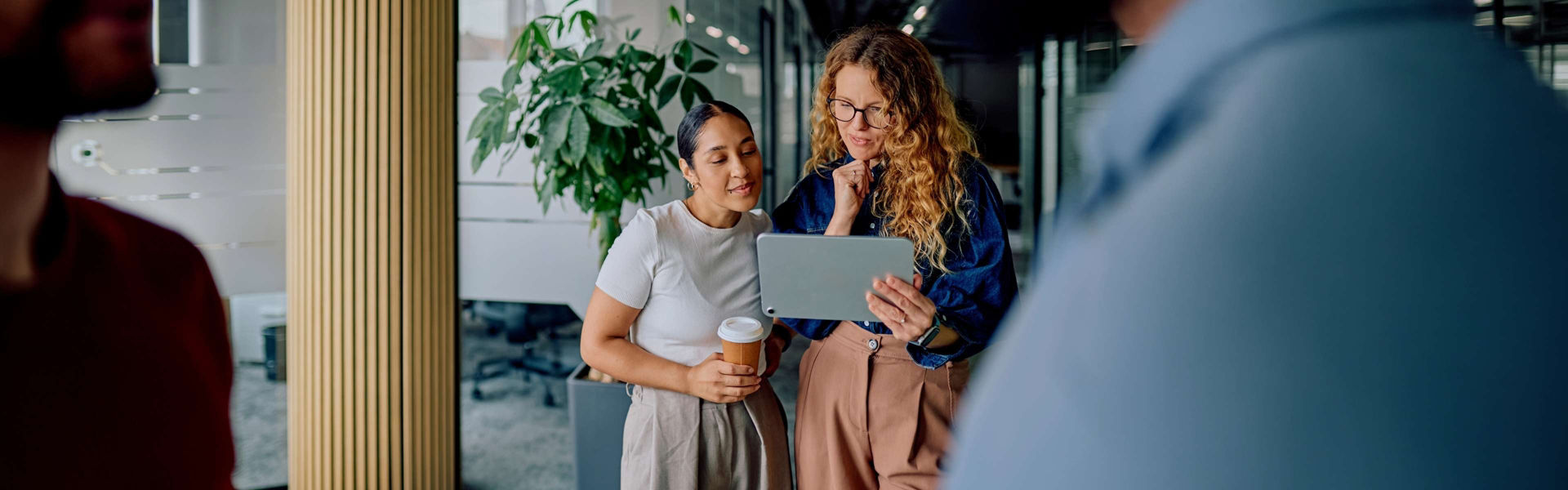 Two women in modern office setting looking at tablet having a conversation about the material, with two people in the foreground.