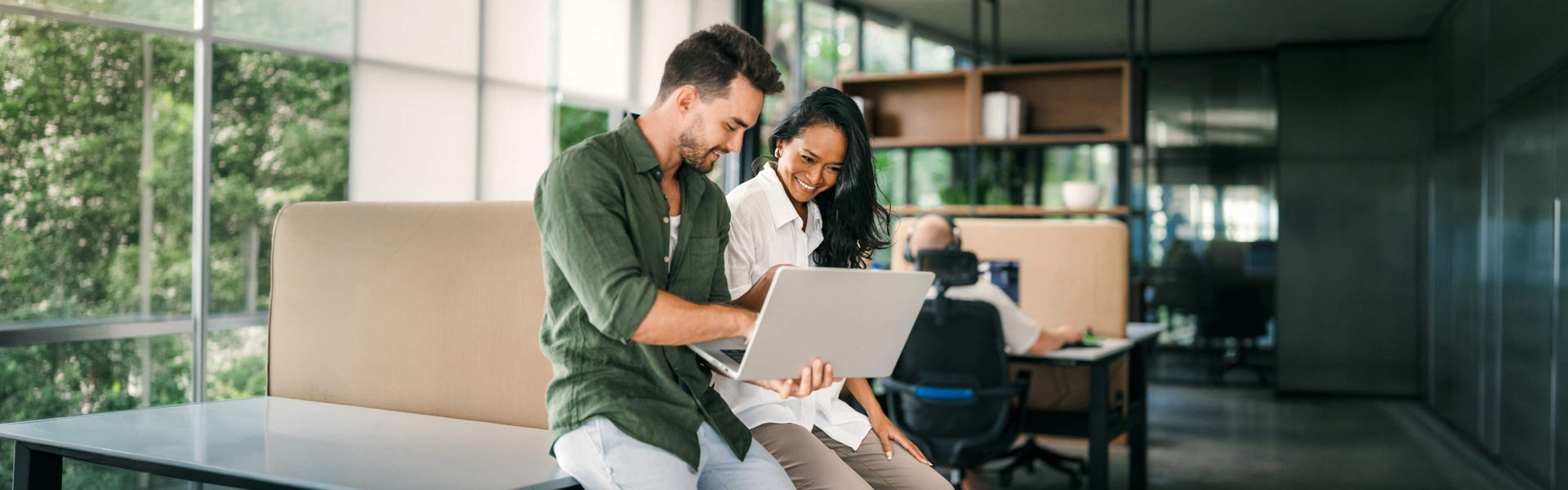 Two colleagues lean on desk in corporate office setting, looking at and discussing something on laptop while smiling wearing business casual outfits