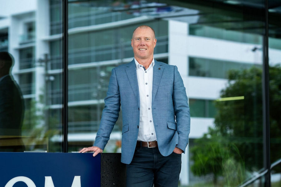 Mid distance shot of Mike Walls standing in front of Datacom Auckland office leaning on Datacom sign, wearing a blue blazer and white shirt, smiling.