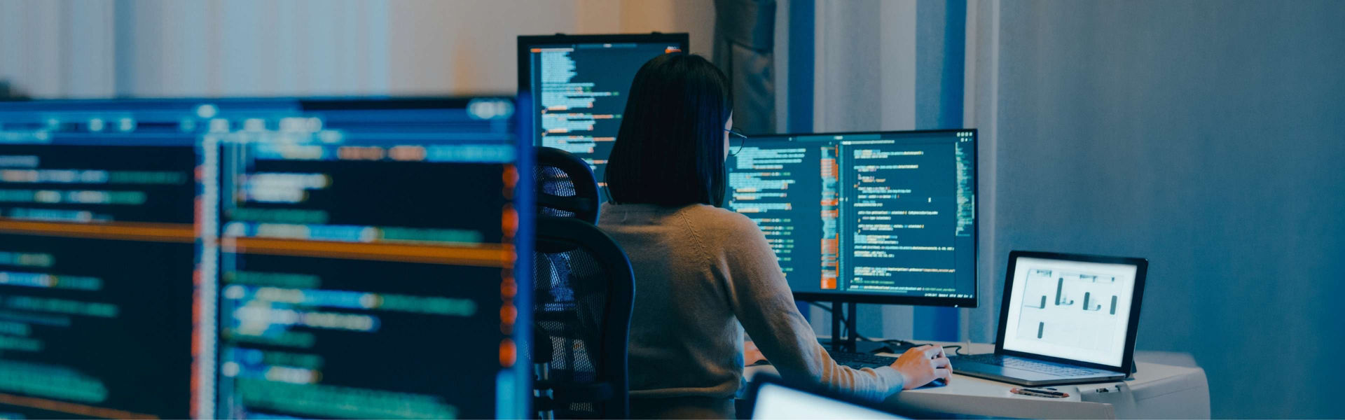 Woman sits at desk in office setting working with computer screens indicating data.