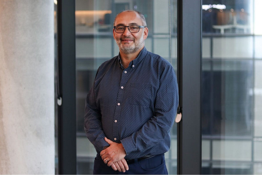 Rich Williams, GM of Data and Analytics at Datacom, stands in front of an office window in a mid-distance shot with his hands resting in front of him, wearing glasses and a blue shirt.