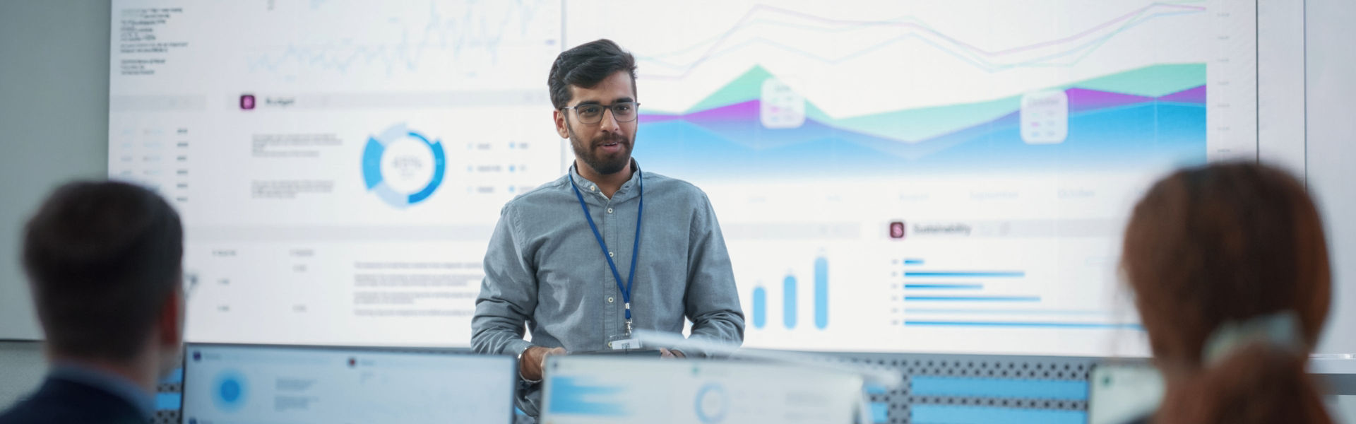 Man presenting data findings in office setting, with data visualisation behind him and the back of audience heads and screens blurred in foreground. He is wearing glasses, blue shirt and lanyard.