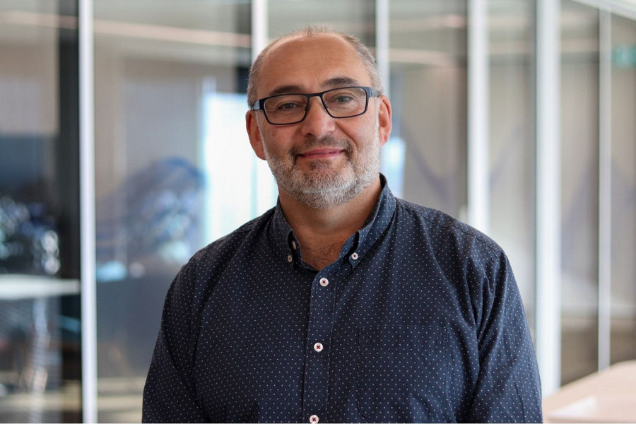 Rich Williams, GM of Data and Analytics at Datacom, stands in front of office meeting rooms, wearing glasses and a blue shirt.