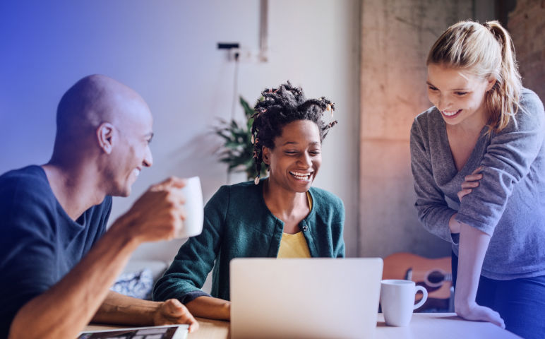 three co-workers sitting around a table, drinking coffee and looking at a laptop