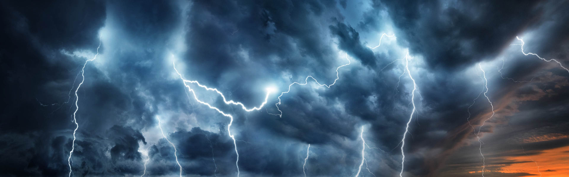 Lightning bolts across dark storm clouds with a hint of sunset on the horizon.