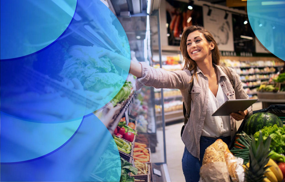 Woman holding an iPad with one hand and reaching out at some vegetables while shopping at the supermarket