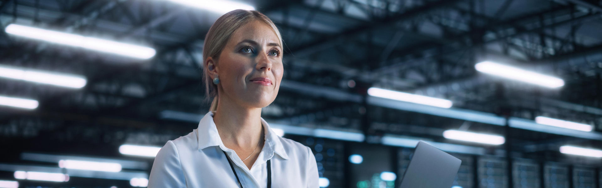 Professional in a white collared shirt with a lanyard holds a laptop inside a large, well-lit industrial facility with metal trusses and bright overhead lights.