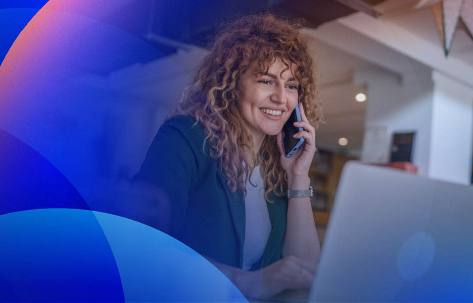 A woman smiling while speaking to customer service on a smartphone while working on a laptop.