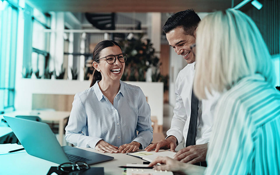 Two women and a man at and office desk in front of a laptop laughing