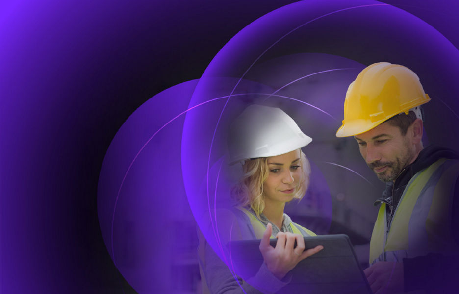 Two individuals looking at an iPad while wearing hard hats and hi-vis vests at a construction site