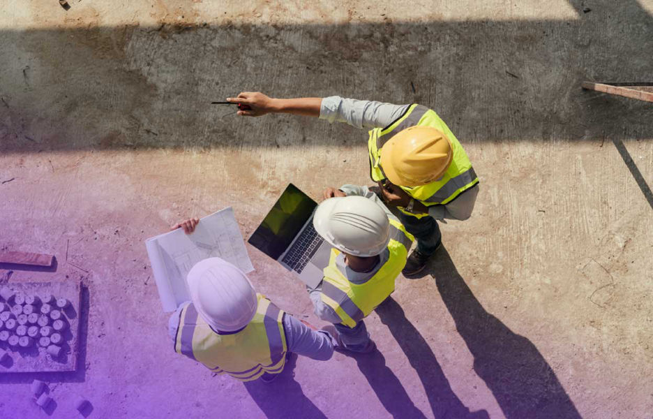 Aerial view of three workers on a construction site holding a paper work and a laptop while looking out on the site