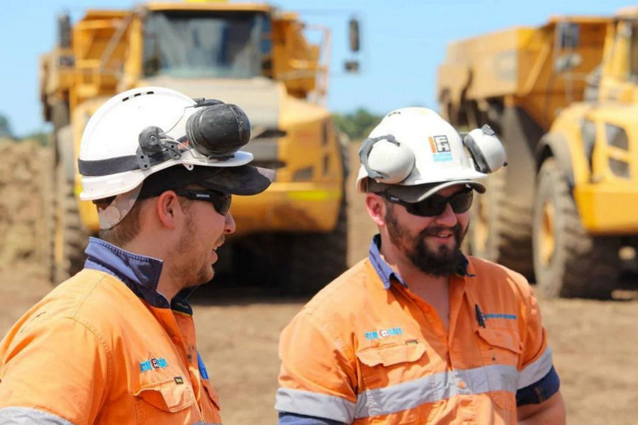 Construction tradespeople wearing orange worksuits, white safety helmets, black sunglasses standing in front of yellow digger and truck on site talking and looking at something off camera