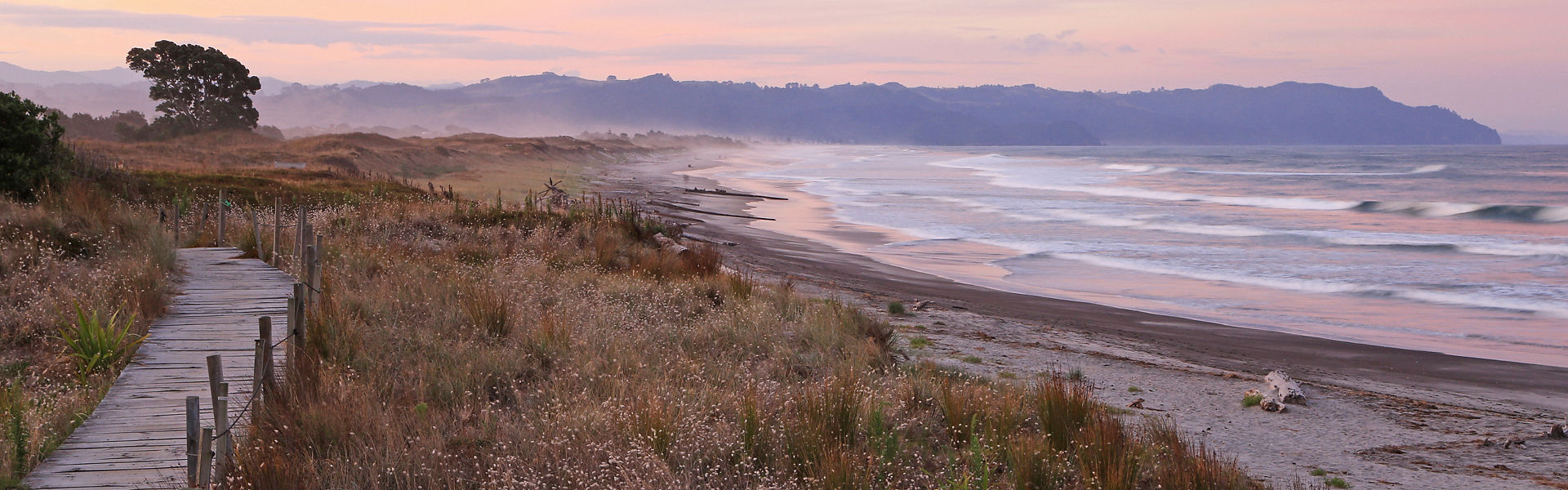 View looking down the length of Waihi beach during pink sunset, path to beach through sand dunes on left, waves and sand on right, hillside in background