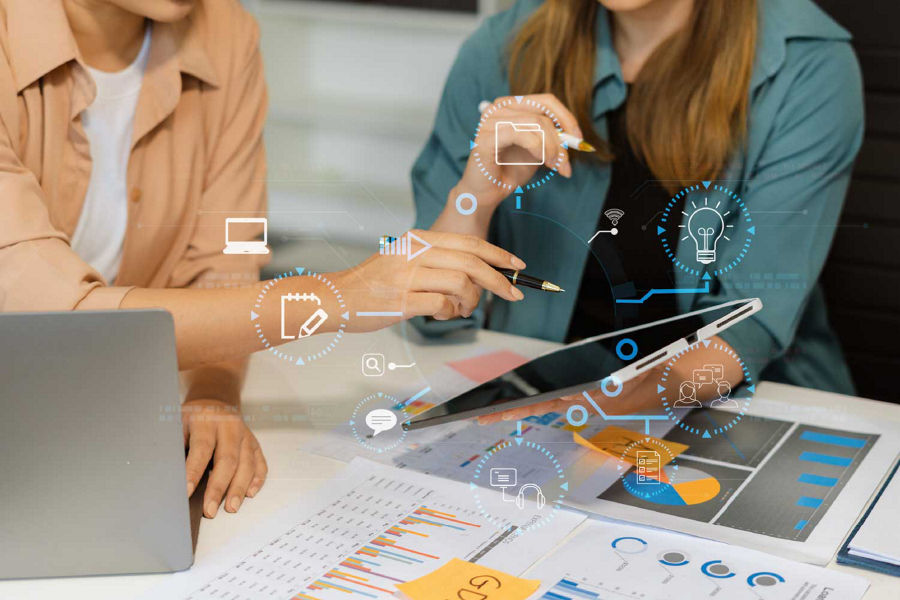 Two individuals collaborate with a tablet at a desk filled with documents