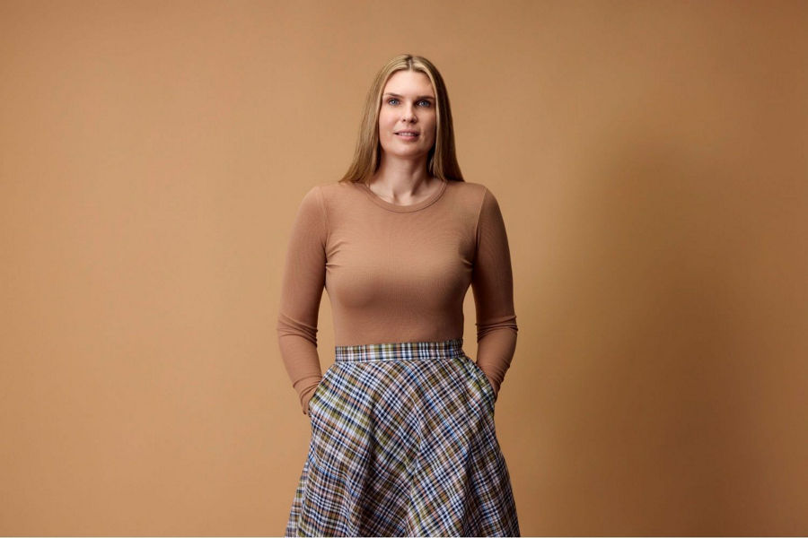 Mid distance shot of Lou Compagnone standing in front of plain background looking at camera with her handsin her pockets, wearing a tan long sleeve and plaid skirt.