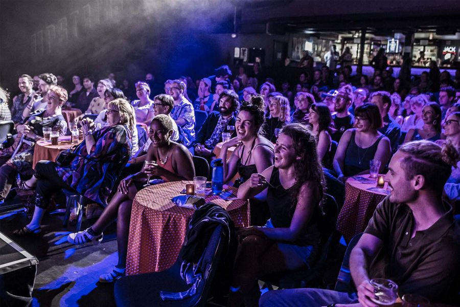 Crowd at a comedy show sitting around tables laughing in purple stage lighting