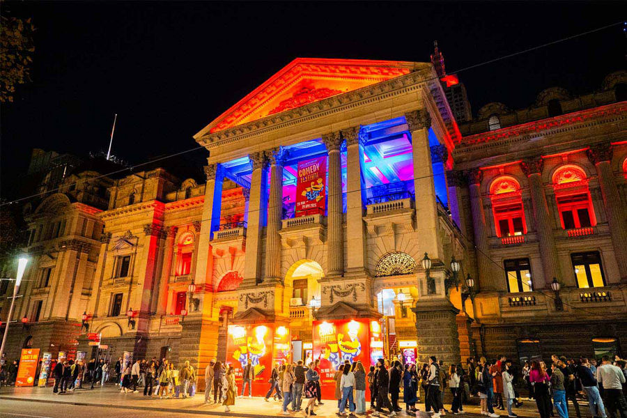 Historical building lit up at night for the Melbourne International Comedy Festival with crowd gathered outside 