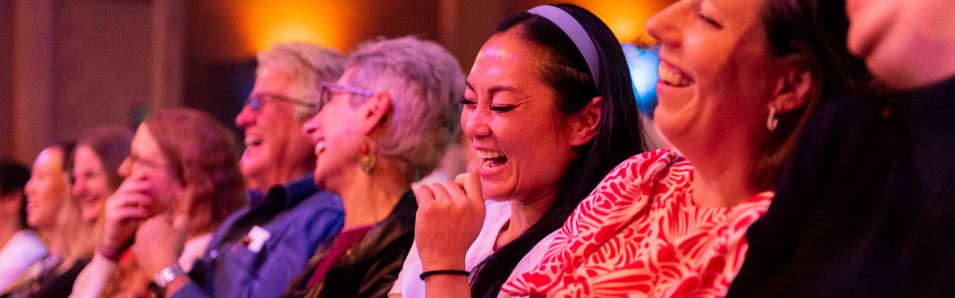 Crowd sitting in a row laughing in a dimly lit indoor setting