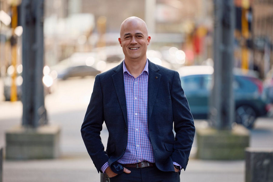 Headshot of Dan Bowbyes wearing blue jacket and checked shirt with hands in pockets standing in Wellington street.