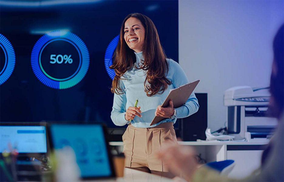 A woman holding a folder standing in front of a screen providing data insights. 