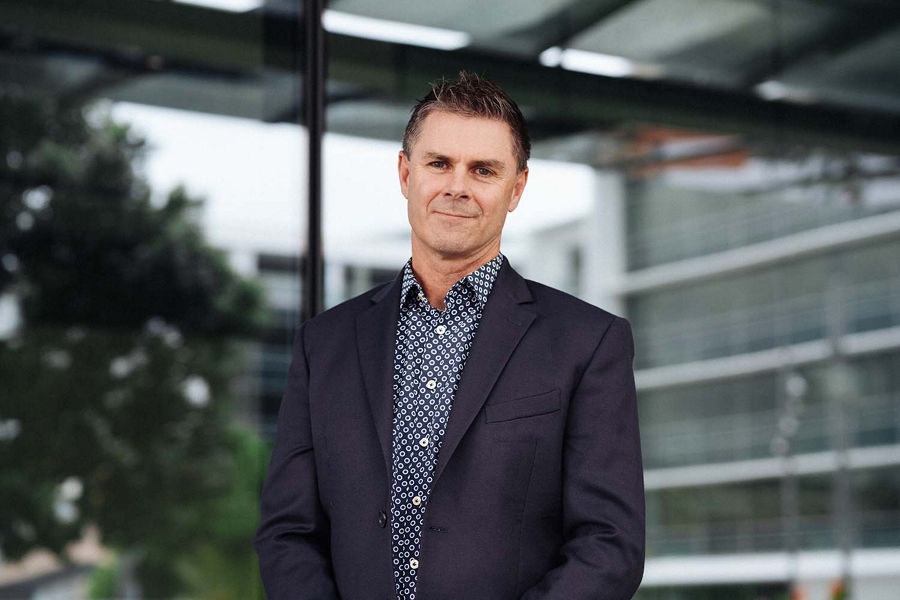 A photo of Peter Nelson standing outside the Auckland office wearing a navy blue suit jacket.