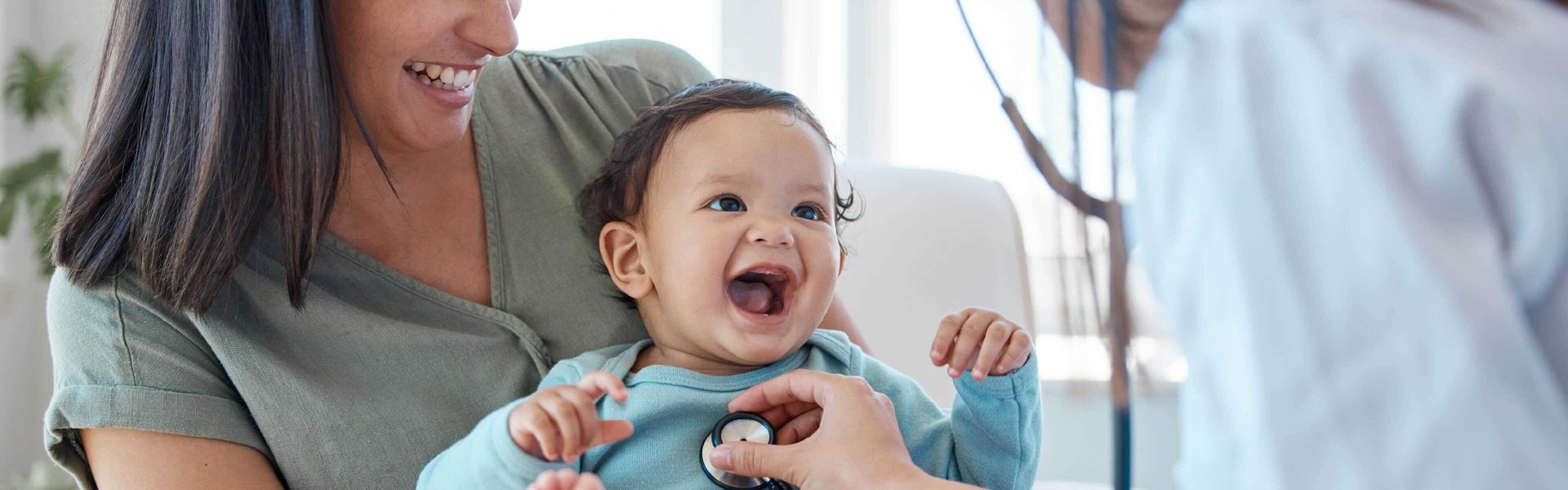 Smiling baby being held by mother while it's heartbeat is being listened to by a doctor through a stethoscope.