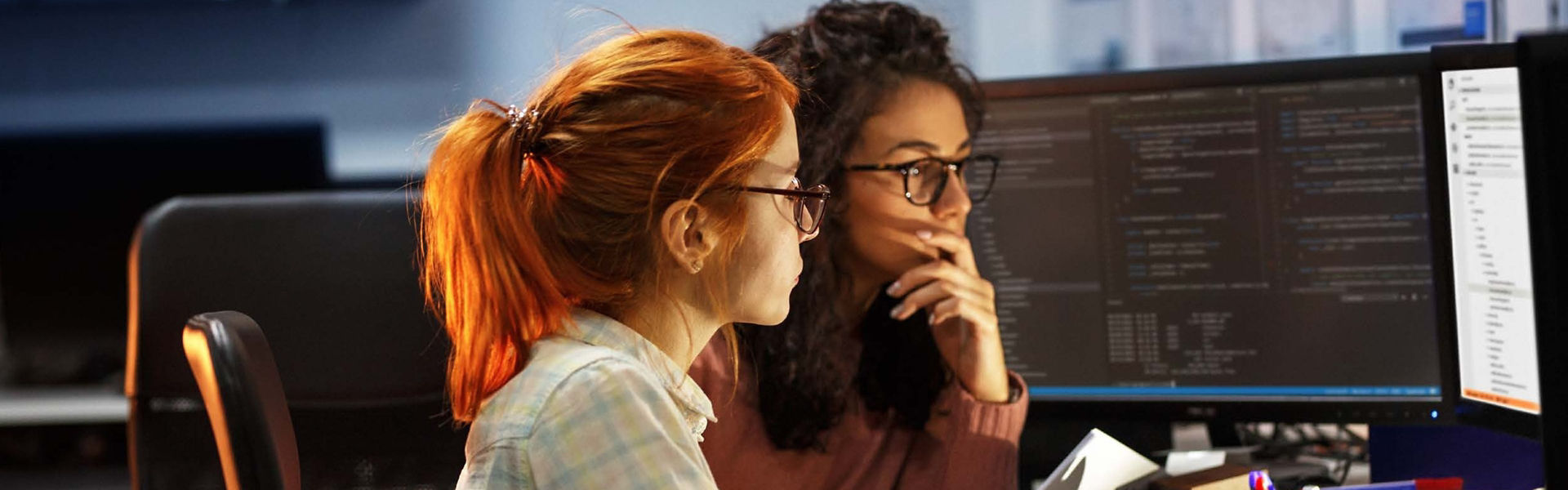 Two women, one with red hair one with dark hair, working at computers together with serious faces 