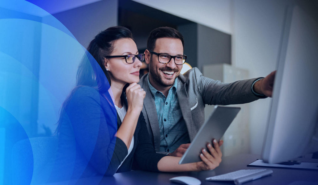 Two business professionals working together, one holding a tablet and the other pointing at a desktop monitor.