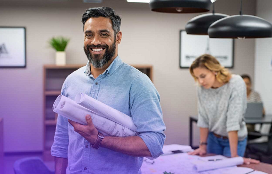 Man in office holding scrolls of paper with women in the background looking at a table full of paperwork