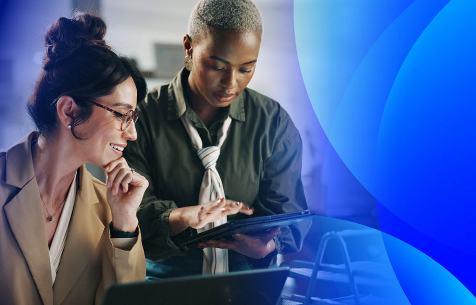 Two women at an office desk interacting with a tablet and laptop.