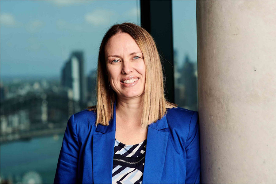 Stacey Tomasoni standing in front of window looking out to city smiling wearing blue blazer with medium length hair