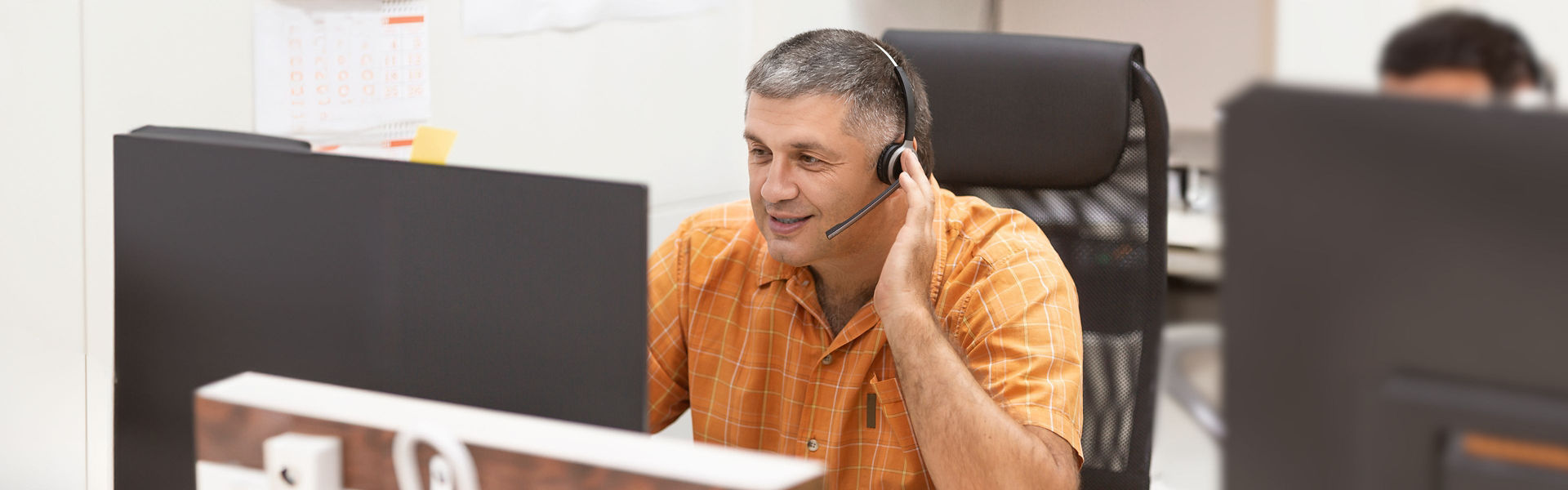Mean wearing headset and orange polo shirt talking to someone while looking at screen in office setting