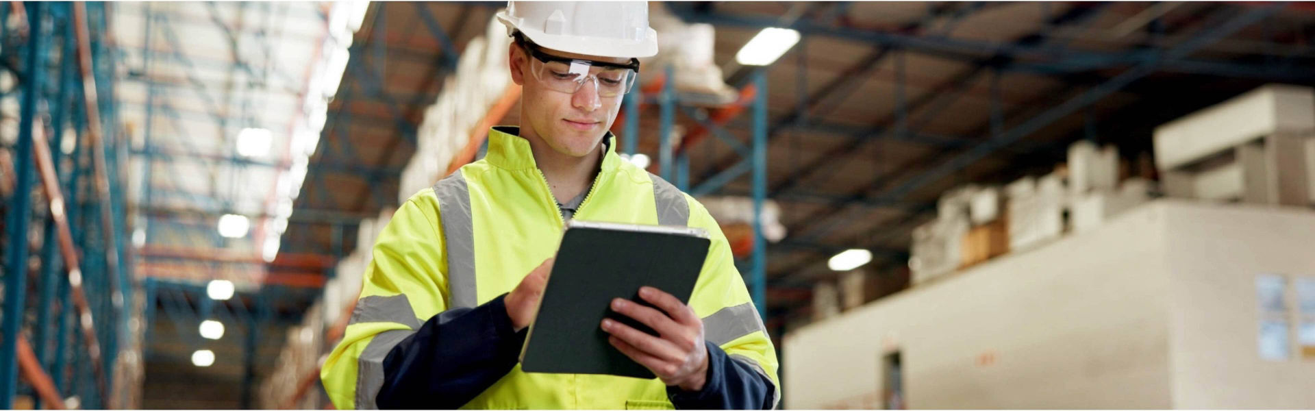 Professional in a white collared shirt with a lanyard holds a laptop inside a large, well-lit industrial facility with metal trusses and bright overhead lights.