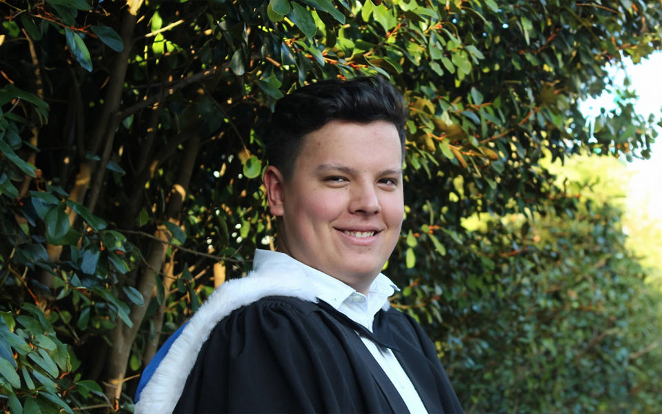 Photograph of Baxter Kirikiri smiling directly at the camera wearing a graduate gown.