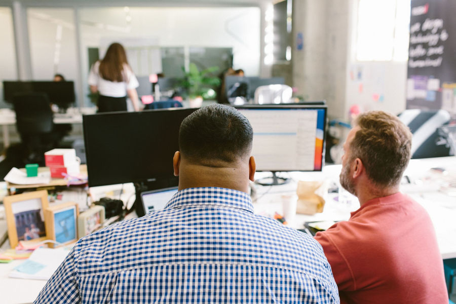 Photo of two men, one wearing a blue checked shirt and the other a salmon pink shirt, looking at a computer screen
