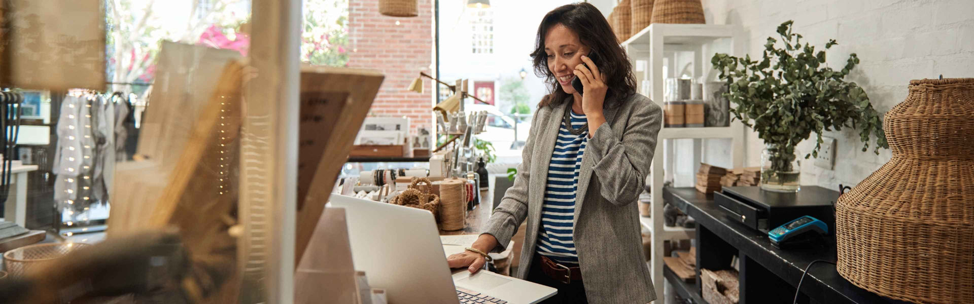 Retail shop owner stands at store counter while speaking on phone and using laptop, wearing blazer and surrounded by wares.