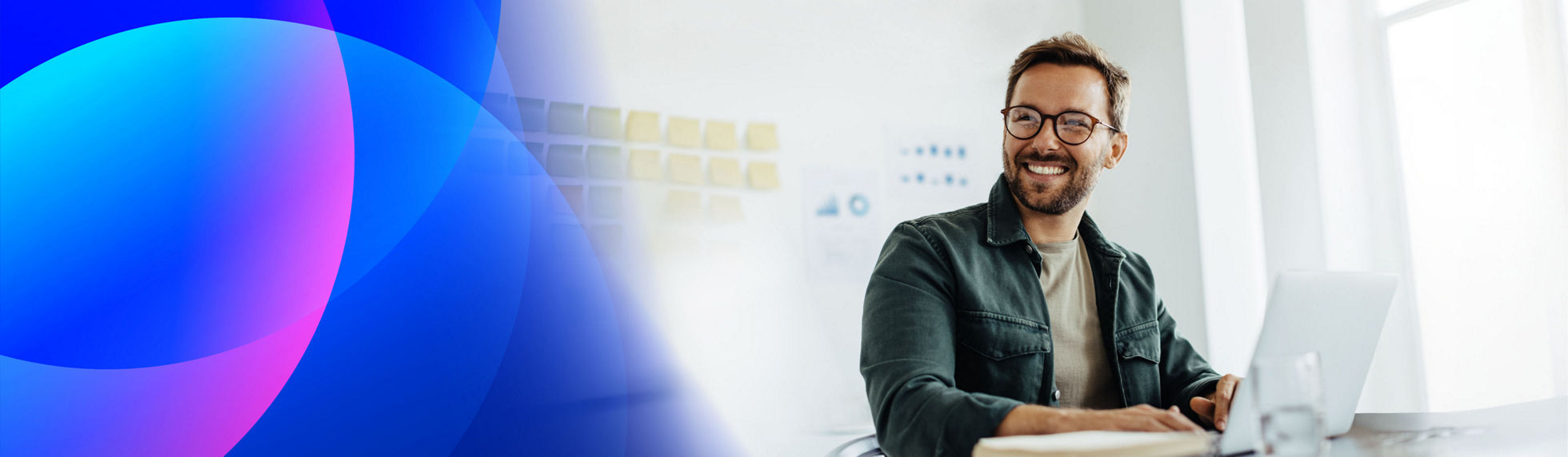 Smiling man in glasses working on a laptop in a bright office, with blue and pink abstract shapes on the left side of the banner.
