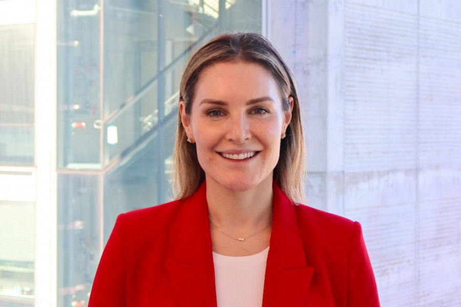 Holly Hunt standing in front of window wearing red blazer, smiling with hair tucked back.