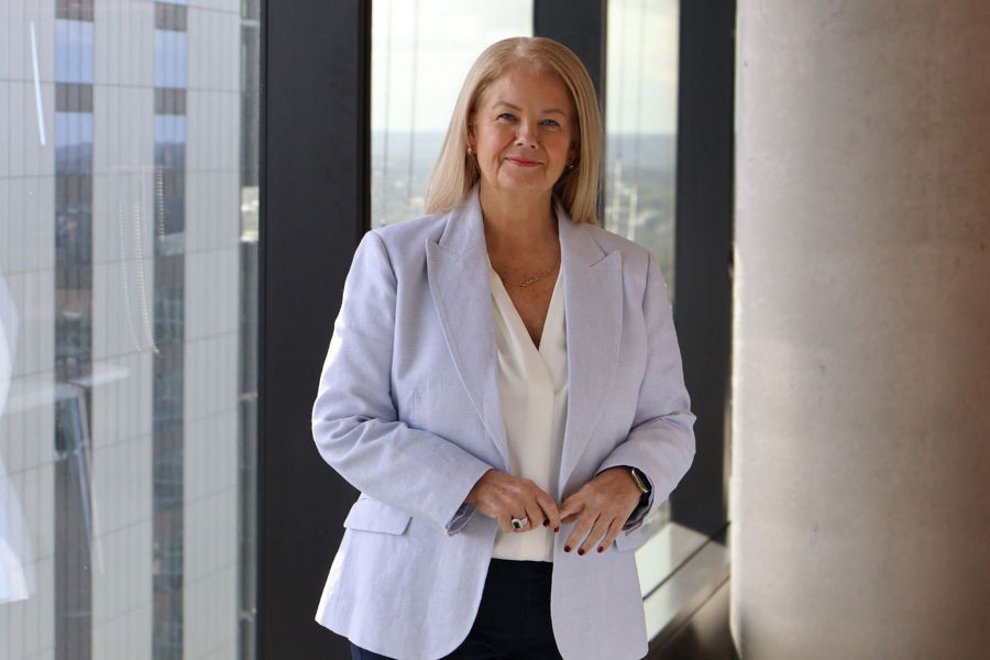 Siobhan stands in front of windows in office building wearing light blue blazer, white shirt. Her hands are in front of her.