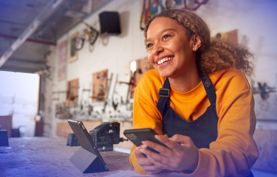 A business owner leaning on a workbench while holding a smartphone next to a tablet on a stand.